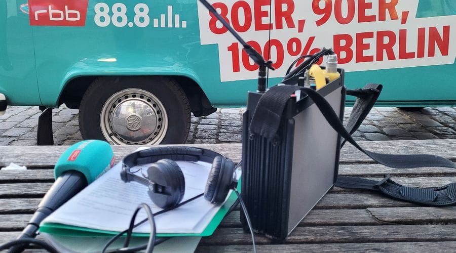 Ein türkisfarbener Lieferwagen mit der Aufschrift "rbb 88.8" und "80ER, 90ER, 100% BERLIN" steht auf Kopfsteinpflaster, davor liegt Radioequipment mit Mikrofon, Kopfhörern und technischen Geräten auf einer Holzbank.