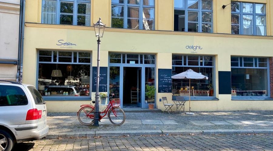 Außenansicht des Cafés Delight in der Bölschestraße Friedrichshagen mit gelber Fassade, blauen Fensterrahmen und Außenbestuhlung. Vor dem Gebäude stehen ein Auto und ein rotes Fahrrad an einem Laternenpfahl auf dem Kopfsteinpflaster.