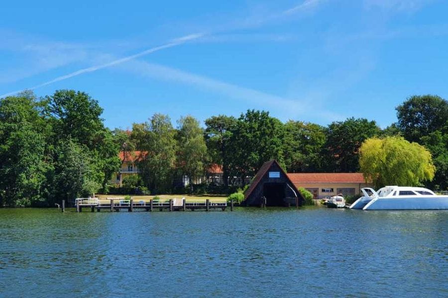 Blick über das Wasser auf das Waldhotel Am See mit roten Ziegeldächern zwischen grünen Bäumen, davor ein Holzsteg und weiße Motorboote am Ufer.