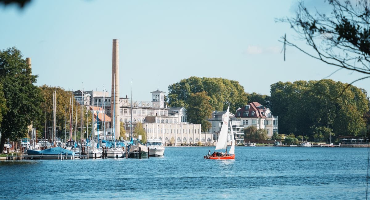Blick auf die Müggelsee Brauerei am Ufer des Müggelsees mit dem charakteristischen hohen Schornstein und den historischen Brauereigebäuden. Im Vordergrund sind ein Segelboot auf dem Wasser und eine Marina mit verschiedenen Booten zu sehen.