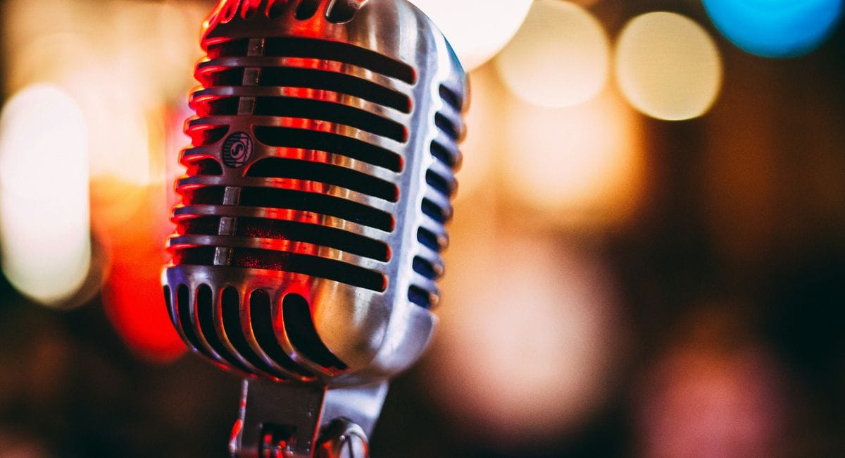 Ein vintage Mikrofon im Retro-Stil mit horizontalen Metallstreifen steht im Vordergrund, während im Hintergrund verschwommene bunte Lichtpunkte in rot, blau und gelb zu sehen sind.