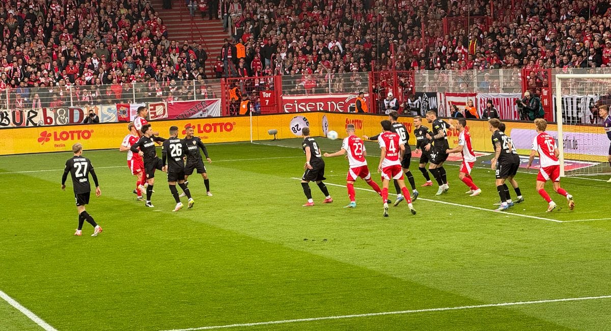 Fußballspiel im Stadion An der Alten Försterei zwischen Union Berlin (rote Trikots) und einer Mannschaft in schwarzen Trikots, mit einer Spielsituation im Strafraum vor einer dicht gefüllten Zuschauertribüne.