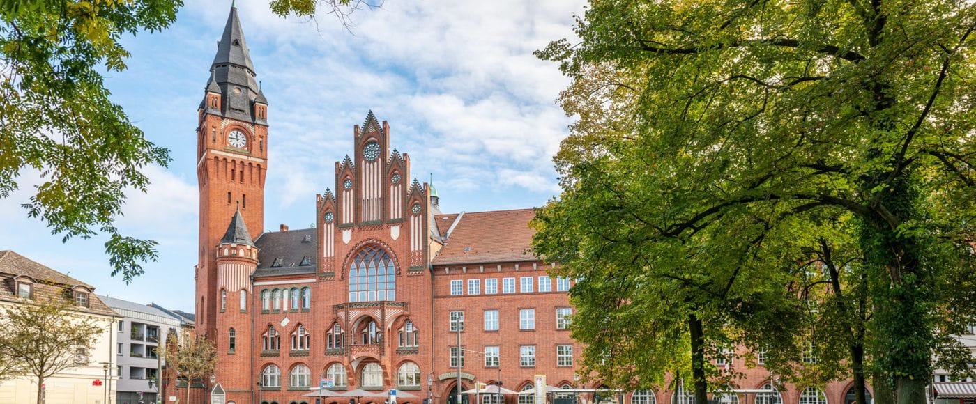 Ein historisches rotes Backsteingebäude mit hohem Uhrenturm und gotischen Architekturelementen steht hinter einer grünen Parkanlage mit großen Laubbäumen. Das Rathaus Köpenick zeigt eine imposante Fassade mit spitzbogigen Fenstern und einem markanten Turm mit Spitzdach.