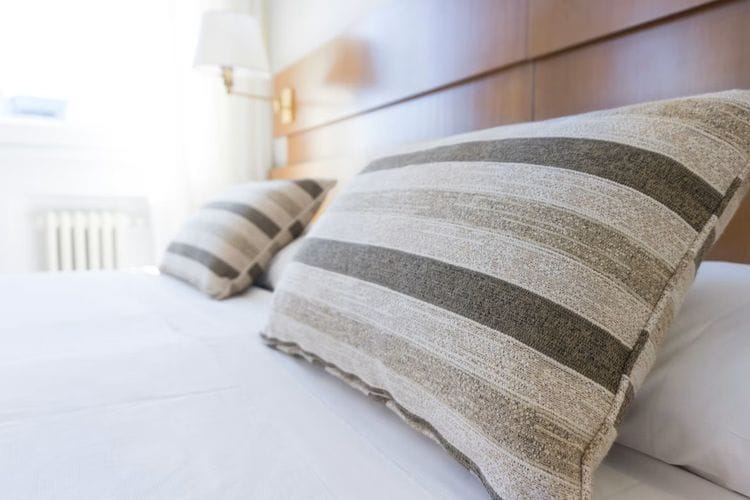 Zwei gestreifte Kissen in Grau- und Beigetönen liegen auf einem weißen Bett vor einem hölzernen Kopfteil. Im Hintergrund ist unscharf eine Lampe und helles Tageslicht durch ein Fenster zu erkennen.