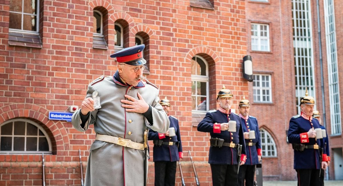 Der Hauptmann von Köpenick in grauer historischer Militäruniform mit roter Mütze steht vor einer Gruppe von Männern in blauen Uniformen mit Gewehren vor einem roten Backsteingebäude. Die Szene zeigt eine historische Militärdarstellung oder Zeremonie auf einem gepflasterten Platz.